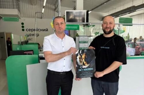 Two men stand at a service center counter, holding a space-themed poster between them and looking at the camera.