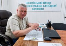 Man seated at a wooden desk in a medical center, looking at the camera with papers and a laptop in front of him; a Ukrainian medical center sign is on the wall behind him.