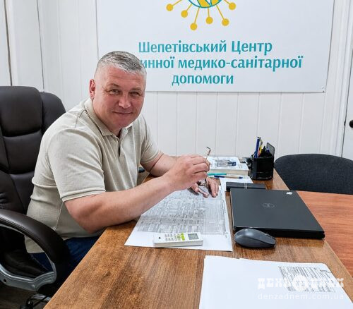 Man seated at a wooden desk in a medical center, looking at the camera with papers and a laptop in front of him; a Ukrainian medical center sign is on the wall behind him.