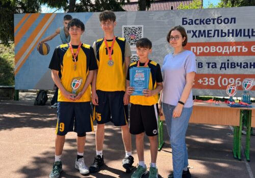 Four young basketball players in yellow and black uniforms with medals, posing with a woman near an event banner outdoors.