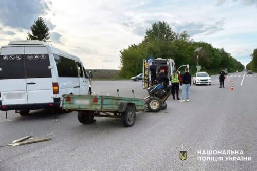 Roadside accident scene with a white van, a small trailer, and an emergency vehicle; bystanders stand on a highway as a traffic cone marks the area, Ukrainian police logo in corner.