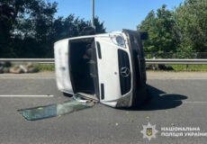White van on its side after a rollover on a highway, shattered glass on the road and debris nearby; police insignia visible in the corner.