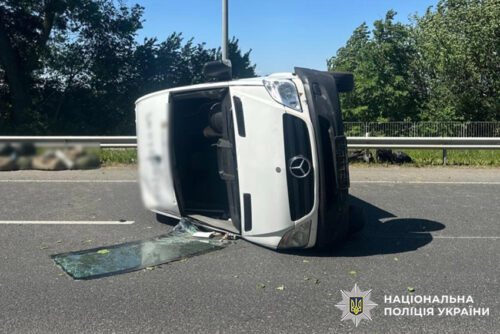 White van on its side after a rollover on a highway, shattered glass on the road and debris nearby; police insignia visible in the corner.