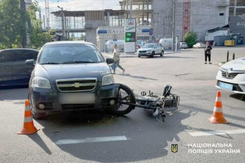 Gray SUV and a fallen motorcycle at an intersection, surrounded by traffic cones; construction site in the background and Ukrainian police logo.