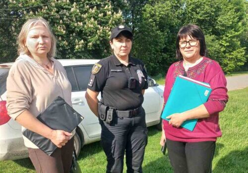 Three women standing on a lawn beside a light-colored car; a police officer in uniform stands between two civilians, each holding folders.