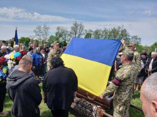 Crowd and uniformed soldiers surround a coffin draped in a blue-and-yellow Ukrainian flag at an outdoor funeral.