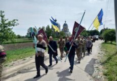 Group of people marching along a rural road, some in camouflage, carrying Ukrainian flags and banners with a church visible in the background.