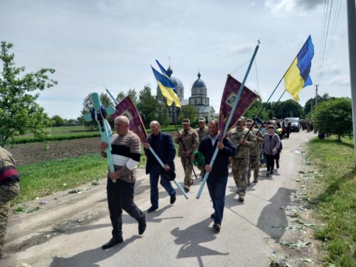 Group of people marching along a rural road, some in camouflage, carrying Ukrainian flags and banners with a church visible in the background.