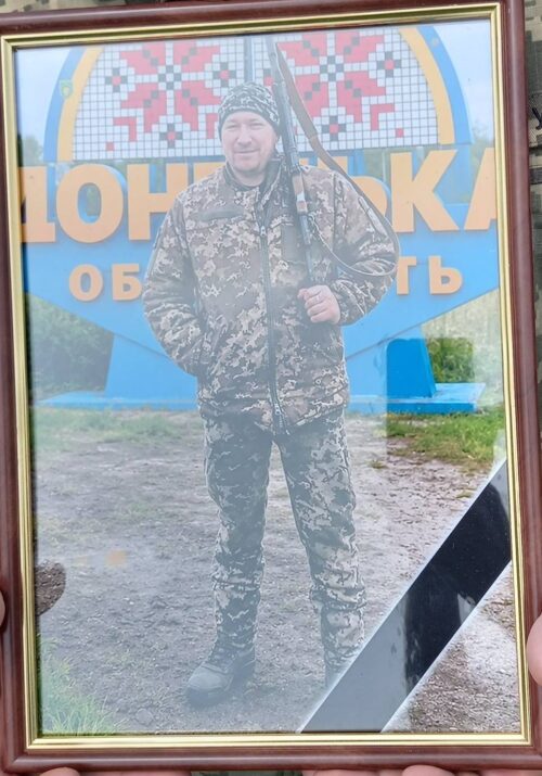 Framed photo of a man in camouflage holding a rifle, standing outdoors with a colorful sign behind him.