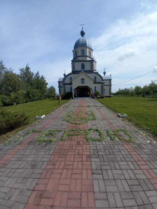 Blue-domed church at the end of a brick walkway, with grass and plants spelling words along the path. core elements: church, dome, entrance, clear day.