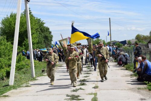 Military personnel marching down a rural road carrying a Ukrainian flag, civilians watching from the roadside on a sunny day.