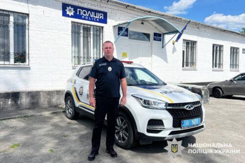 Policeman in uniform standing beside a white police SUV outside a Ukrainian police station, with police signage visible.