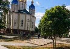 Orthodox church with blue domes in a park, with people cycling nearby.