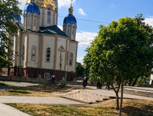 Orthodox church with blue domes in a park, with people cycling nearby.