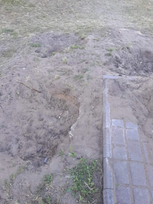 Dirt area with a shallow trench beside a small concrete-tiled border or step, likely a landscaping or foundation excavation.