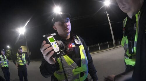 Nighttime roadside police stop: officer in a reflective vest holds a breathalyzer toward the camera, with other officers in the background.