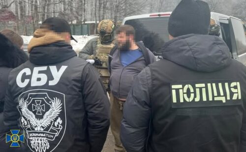 Two officers with SBU and police patches stand behind a man in a blue jacket during a snowy arrest in wintertime outdoors.