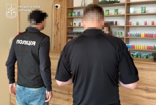 Two men with blurred faces stand in a shop; one wears a jacket labeled 'ПОЛІЦІЯ' and a Ukrainian Prosecutor's Office watermark appears in the corner, shelves visible behind them.