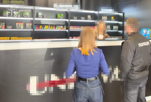 A woman and a police officer stand at a public service counter in a government office, with the Ukrainian Prosecutor's Office logo in the upper left.