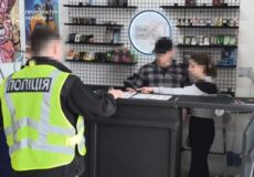 Ukrainian police officer in a high‑visibility jacket labeled 'ПОЛІЦІЯ' talks to two blurred customers at a shop counter inside a store, with shelves in the background and a prosecutor's watermark in the top-left corner.