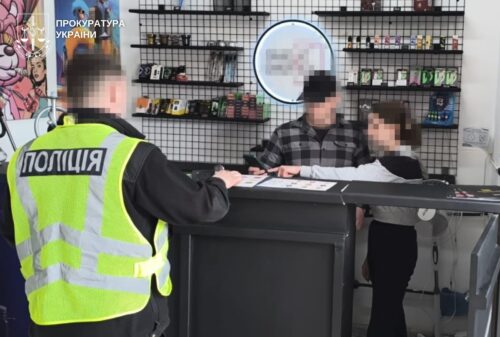 Ukrainian police officer in a high‑visibility jacket labeled 'ПОЛІЦІЯ' talks to two blurred customers at a shop counter inside a store, with shelves in the background and a prosecutor's watermark in the top-left corner.