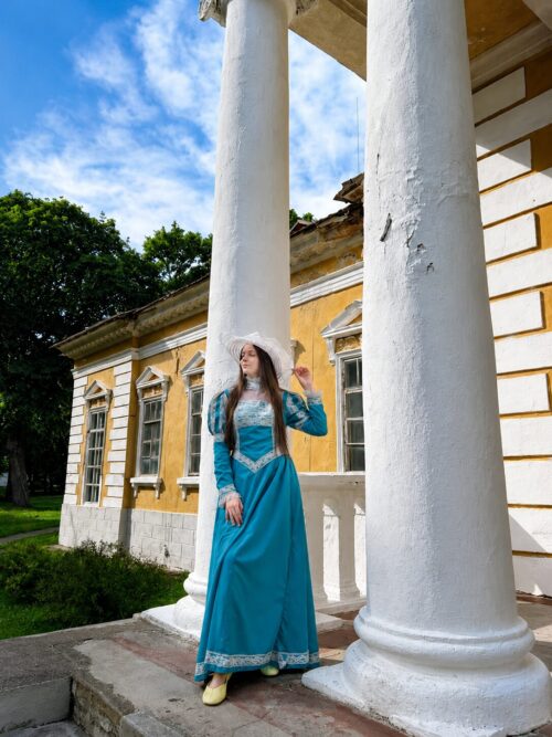 Woman in a blue period dress and bonnet stands on a stone porch between tall white columns of a yellow historic building.