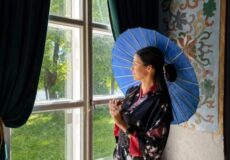 Woman in a dark floral kimono holding a blue parasol, standing by a large window with green curtains and an ornate wall mural to the side.