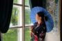 Woman in a dark floral kimono holding a blue parasol, standing by a large window with green curtains and an ornate wall mural to the side.