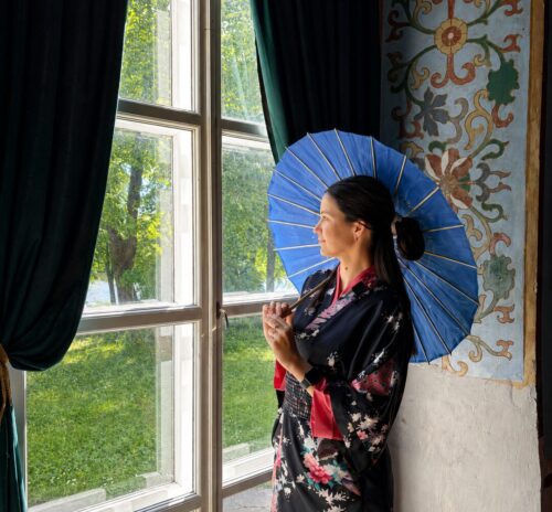 Woman in a dark floral kimono holding a blue parasol, standing by a large window with green curtains and an ornate wall mural to the side.