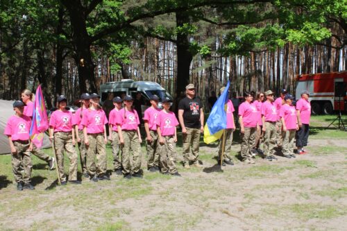 Group of youths in pink shirts and khaki pants stand in a line outdoors, holding flags, with trees and a fire truck in the background.