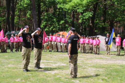 Three soldiers in camouflage and black T-shirts salute on a grassy field during a ceremony, while a line of recruits in pink shirts stands at attention in the background with tents and trees.