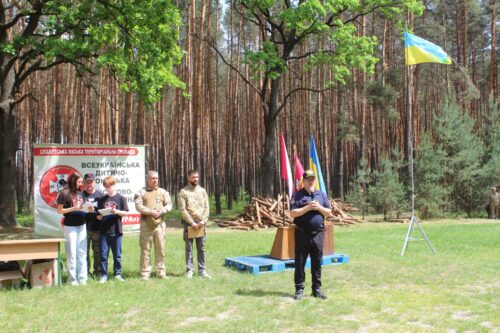 Man speaking into a microphone at an outdoor ceremony in a pine forest; group of people and a banner behind him.