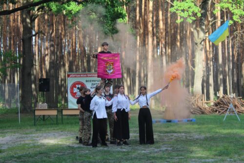 Group of people posing in a forest during an outdoor event, holding a pink banner with a crest, Ukrainian flag nearby, and orange smoke flare in the background.