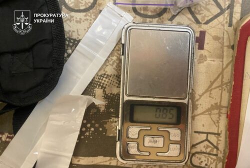 Digital pocket scale shows 0.85 g, with white plastic-wrapped items nearby and a Ukrainian prosecutor's emblem in the corner.