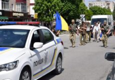 White police car with blue-yellow stripes in the foreground as a group of soldiers marches down a city street during a parade and bystanders watch the scene.