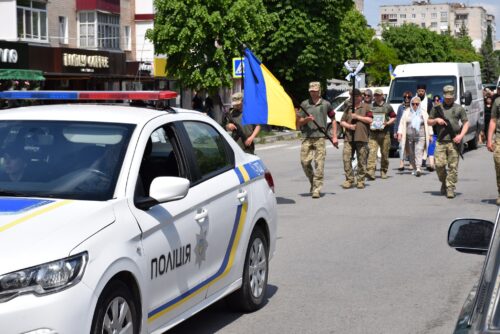 White police car with blue-yellow stripes in the foreground as a group of soldiers marches down a city street during a parade and bystanders watch the scene.