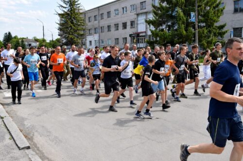 Crowd of diverse runners at the start of a street race on a sunny day, with a building and trees in the background.