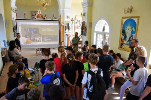 Group of children seated and standing in a church hall watching a presentation on a projector screen in front of them with an adult facilitator nearby.