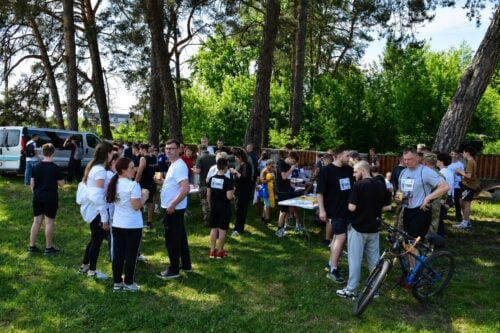 Group of people in a park gathered around tables for an outdoor event, some wearing numbered badges; a bicycle rests near a man, with a van in the background.