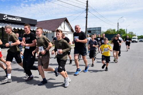 Group of runners in a street race, front runners in green shirts, on a sunny day beside a storefront.