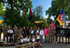 Group of soldiers and civilians posing outdoors with three flags (Ukrainian flag left, red/blue center, another flag right) and certificates in hand.