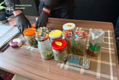 Gloved hands arrange seized jars of cannabis on a table; Ukrainian Prosecutor's Office logo visible in the corner.
