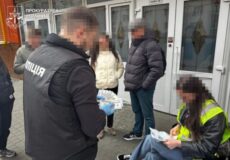 Police officers stand outside a building with blurred civilians; one officer wearing a jacket labeled 'Поліція' holds documents while others watch.