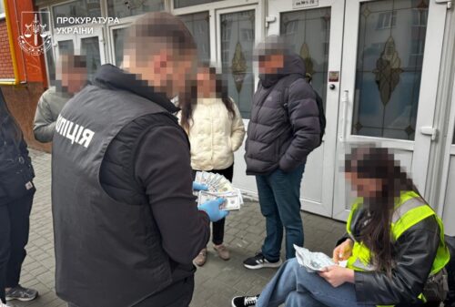 Police officers stand outside a building with blurred civilians; one officer wearing a jacket labeled 'Поліція' holds documents while others watch.