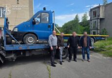 Blue tow truck carrying a blue van on its flatbed; four men stand in front behind yellow tape, Ukrainian Prosecutor’s Office logo in the corner.