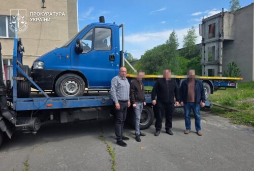 Blue tow truck carrying a blue van on its flatbed; four men stand in front behind yellow tape, Ukrainian Prosecutor’s Office logo in the corner.