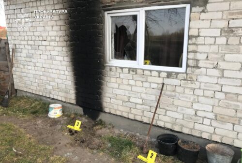 Exterior brick wall with a burned section under the window; a crime-scene setup with yellow evidence markers 1 and 2, plus buckets and tools nearby.