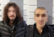 Two blurred adults posing for a police mugshot against a height chart backdrop with measurement lines behind them.