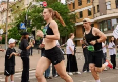 Female runner in black athletic wear leads a street race, with a male runner beside her and spectators along a city sidewalk behind them.
