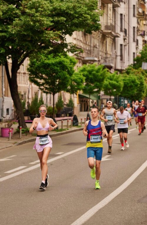 Runners participate in a tree-lined city street marathon, led by a woman in pink sportswear and a man in blue shorts carrying bib numbers.